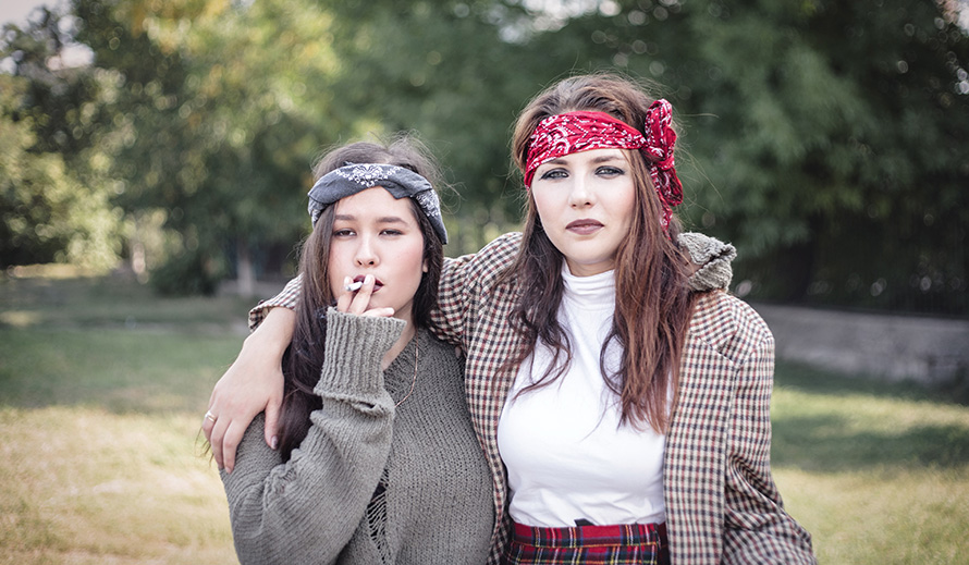 Two informally dressed girls walking down the street with cigarettes.